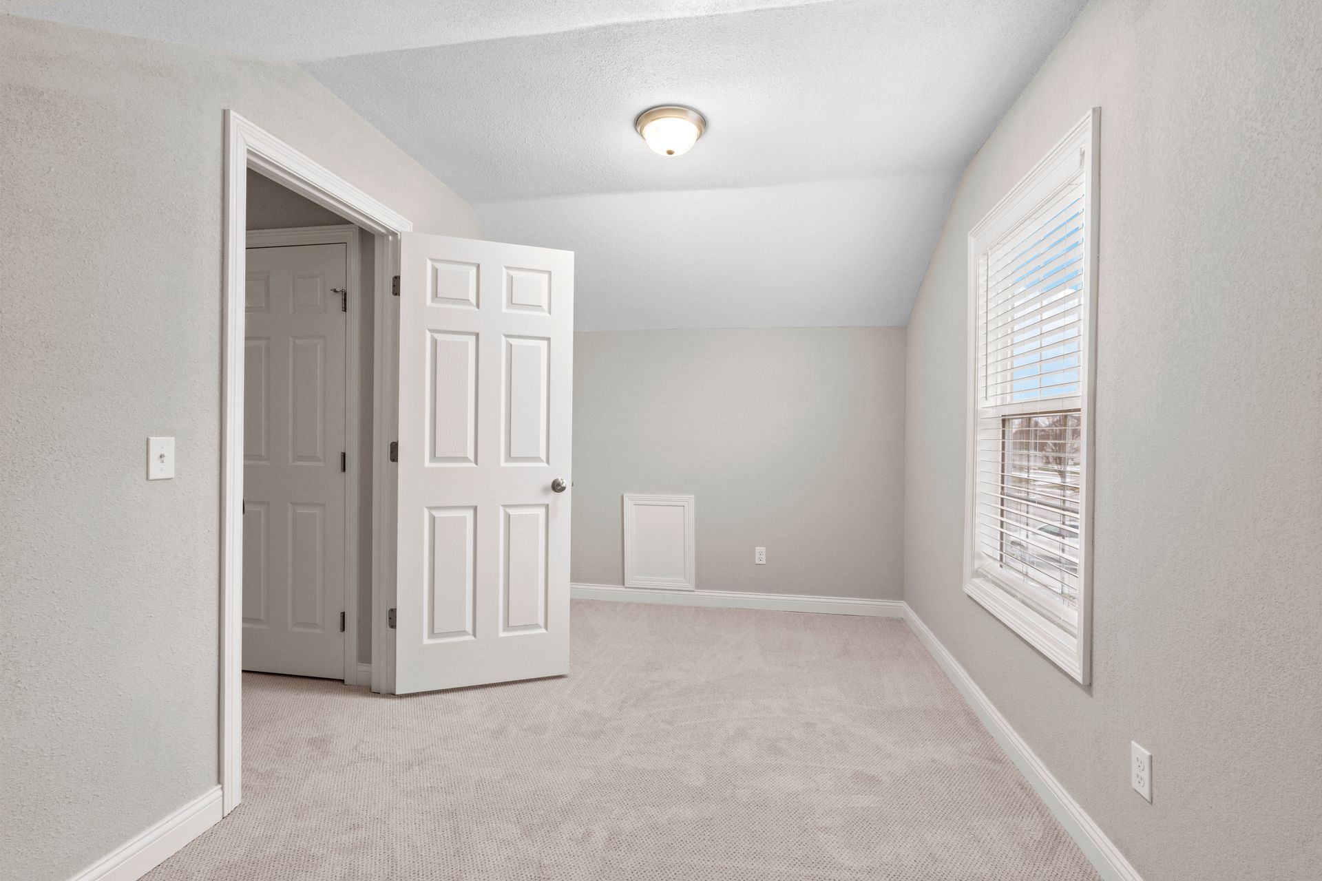 Empty bedroom with light gray walls, white trim, and beige carpet. Window with blinds.