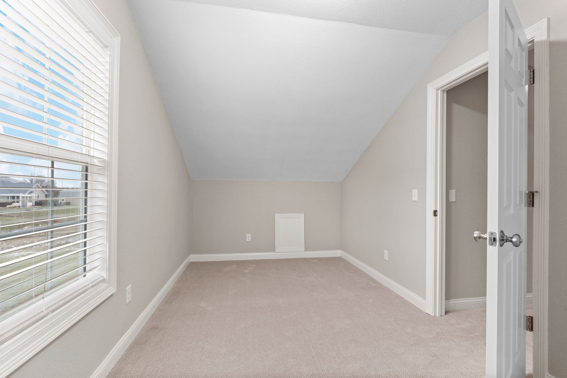 Empty room with sloped ceiling, window with blinds, door, and carpet.