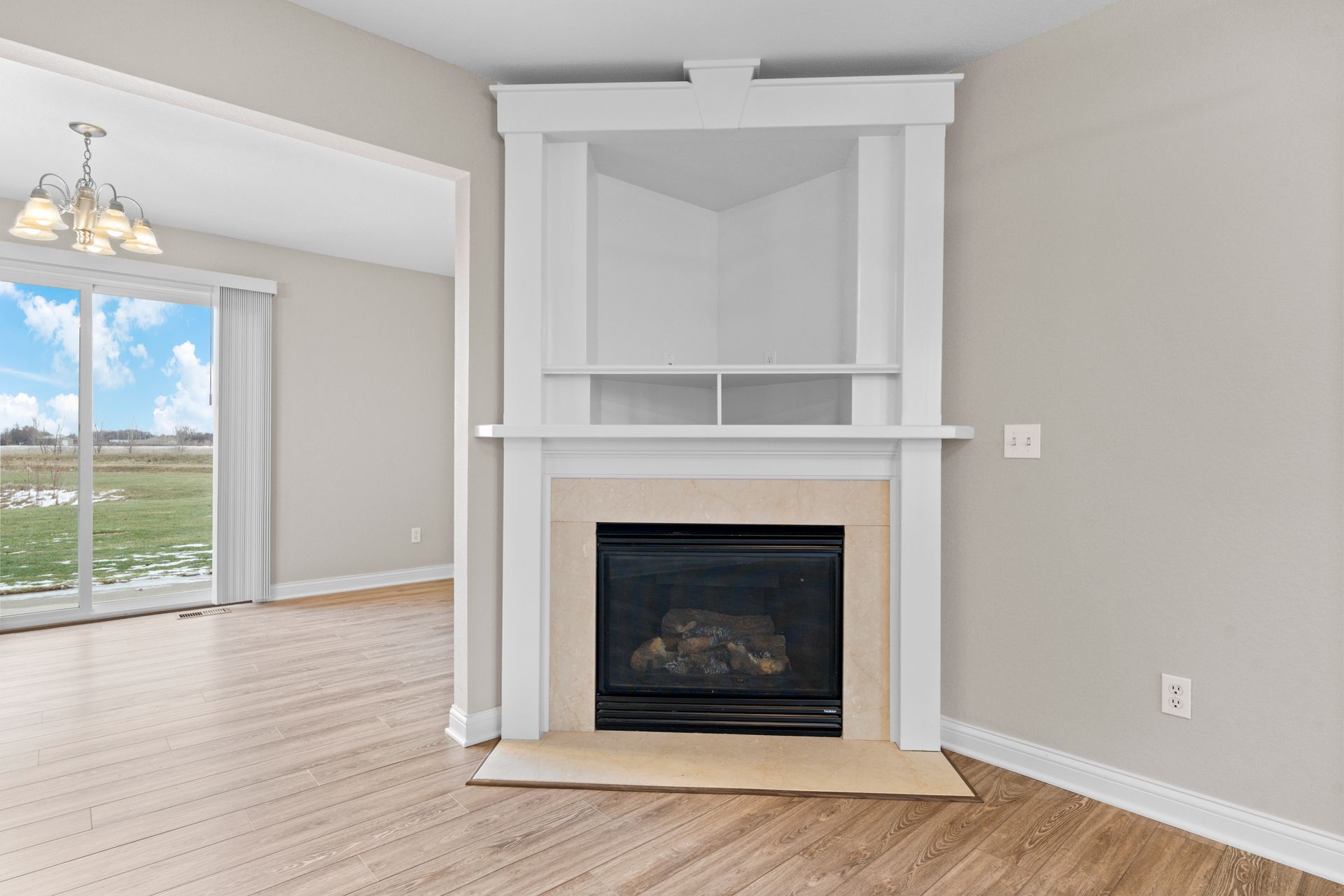 Empty living room with fireplace, white mantle, light gray walls, wood floors, and a view of outdoor landscape.