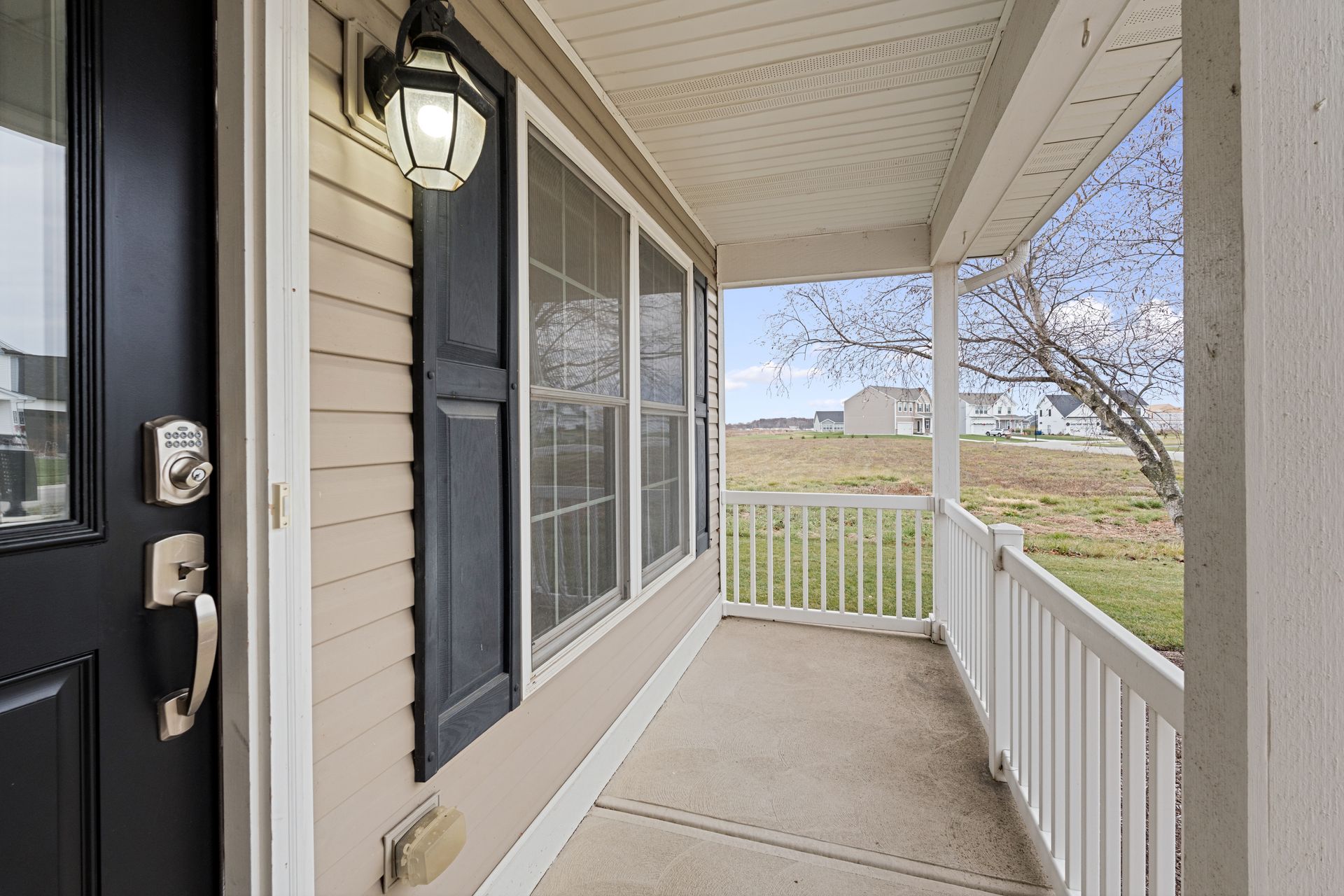 Porch of a beige house with a black door, shutters, and a white railing. The porch overlooks a grassy field.