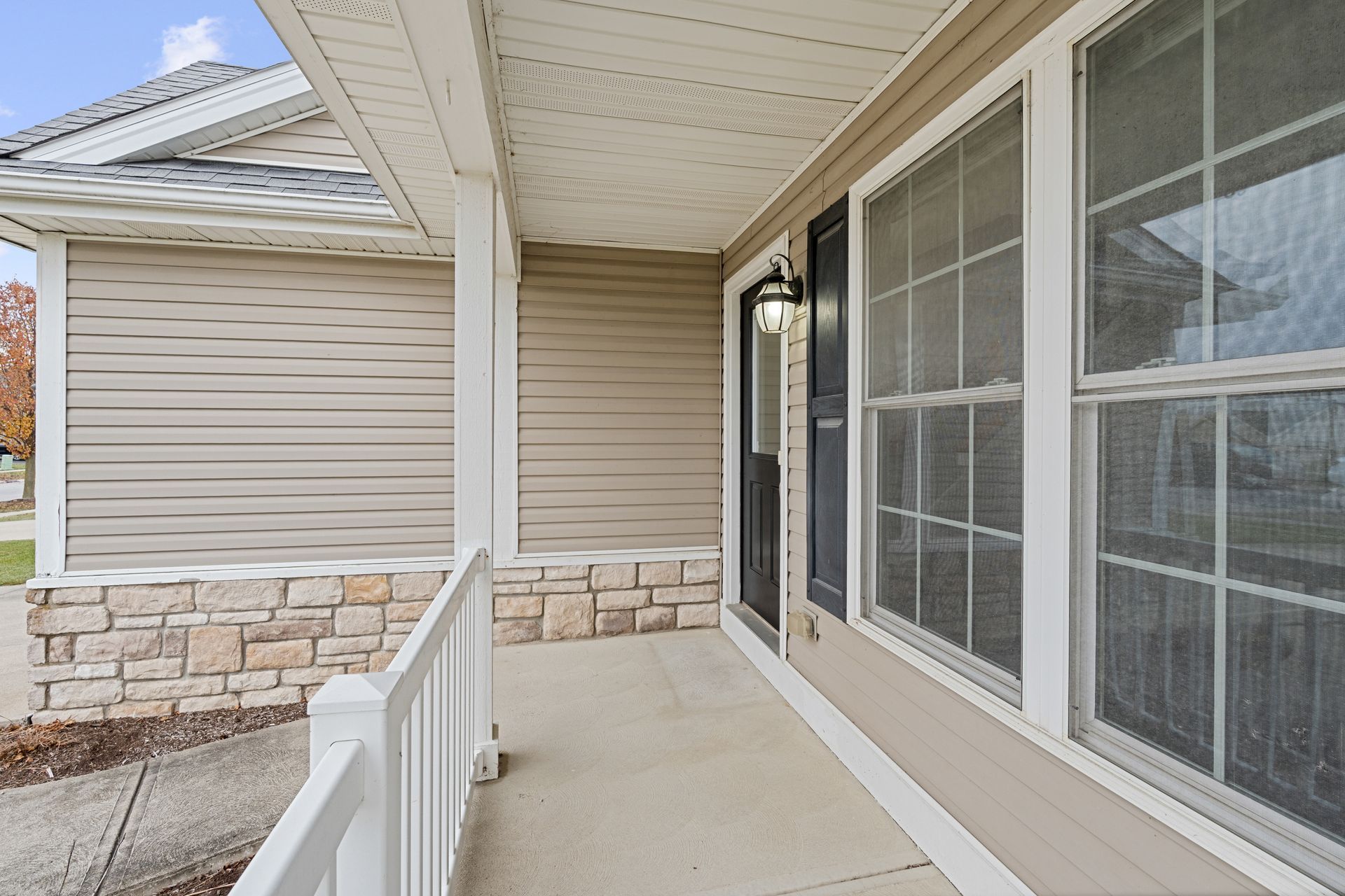 Covered porch of a house with windows, front door, white railing, and stone facade.