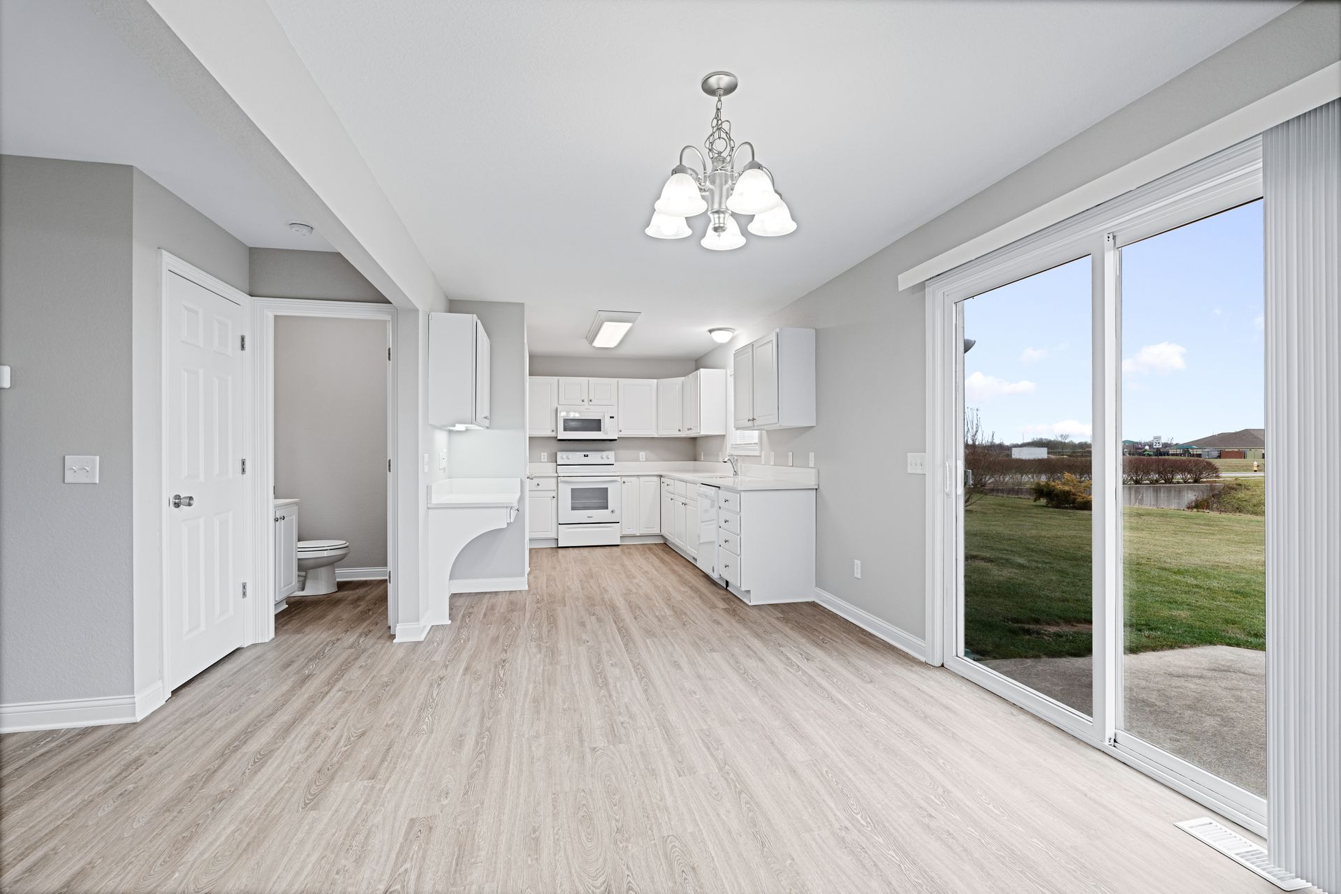 Empty, bright kitchen with white cabinets, appliances, and walls, light wood flooring, and a large sliding glass door.
