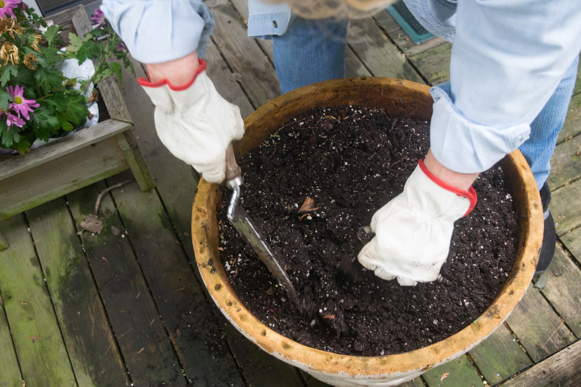 Person in Gloves Uses a Trowel to Mix Soil in a Large Pot on a Wooden Deck — Laurieton Landscape Supplies In Laurieton, NSW