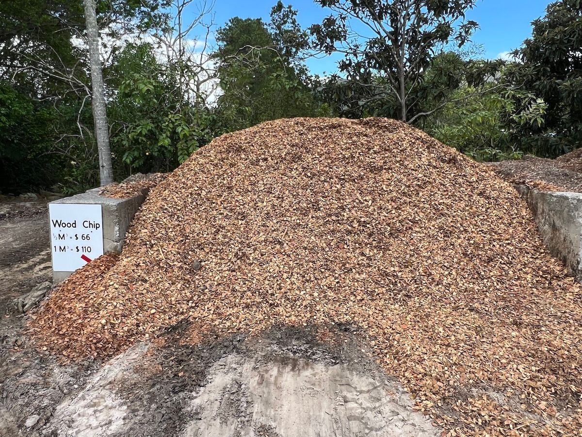 A large pile of wood mulch sits between two concrete barriers outdoors, surrounded by trees and a clear blue sky — Laurieton Landscape Supplies In Lake Cathie, NSW