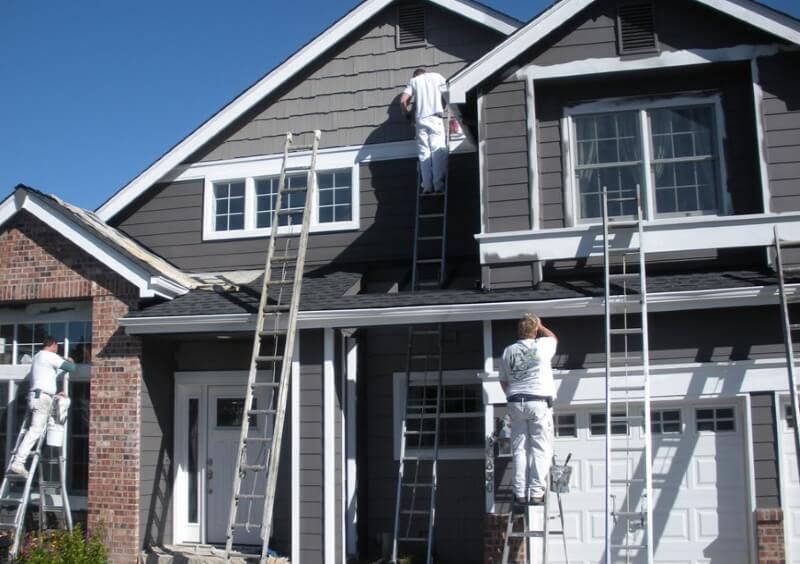 Workers on ladders painting the exterior of a gray house with white trim.
