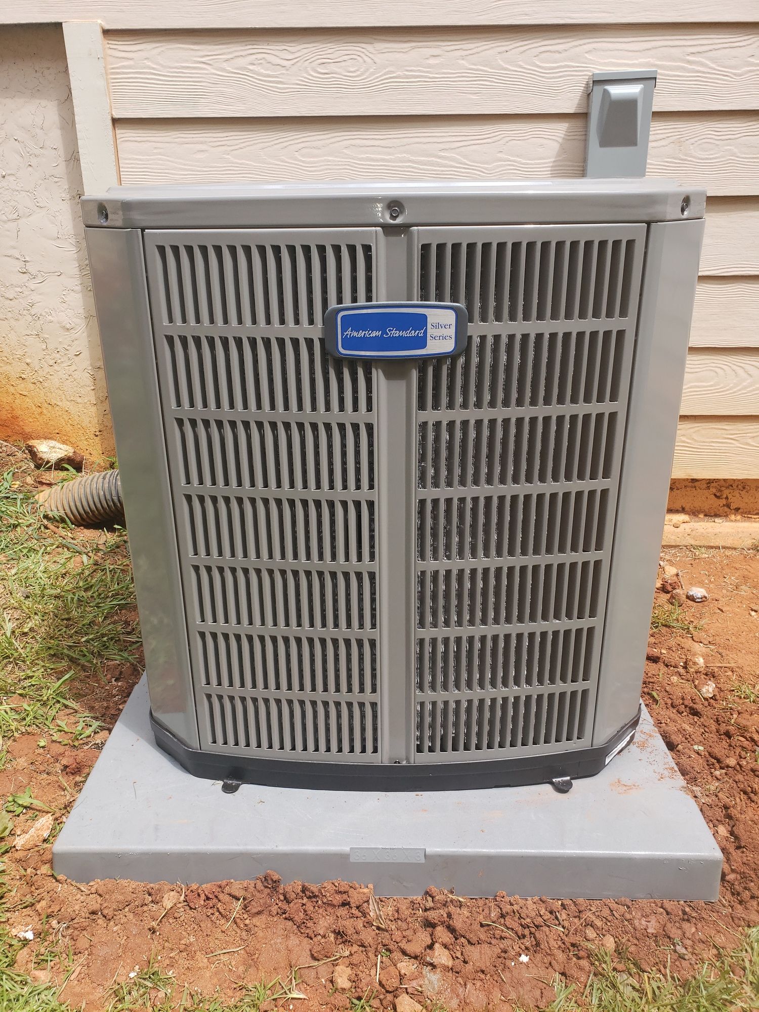A gray air conditioner is sitting on top of a concrete base in front of a house.