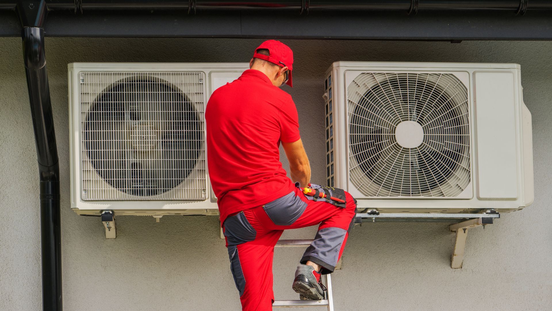 A man is standing on a ladder fixing an air conditioner on the side of a building.