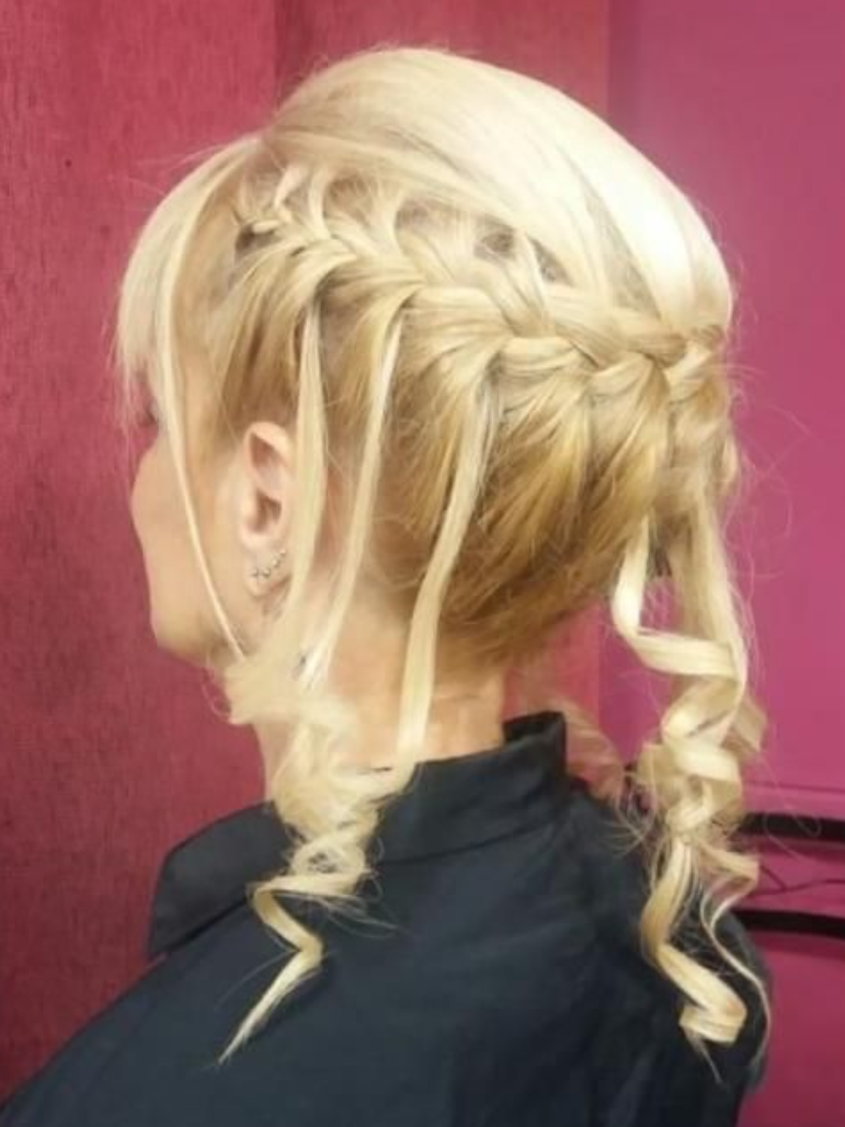Peinado recogido de mujer rubia con corona trenzada y zarcillos rizados, sobre un fondo rosa.