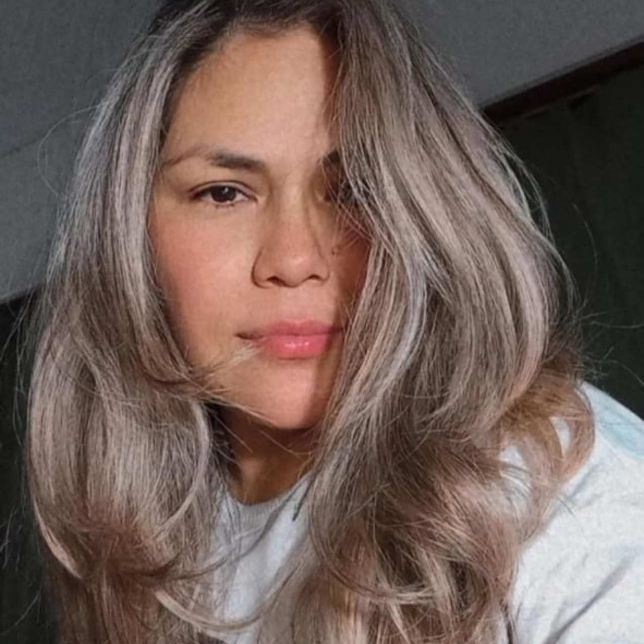 Mujer con cabello gris largo y ondulado y tez clara, mirando a la cámara, vistiendo una blusa de color claro.