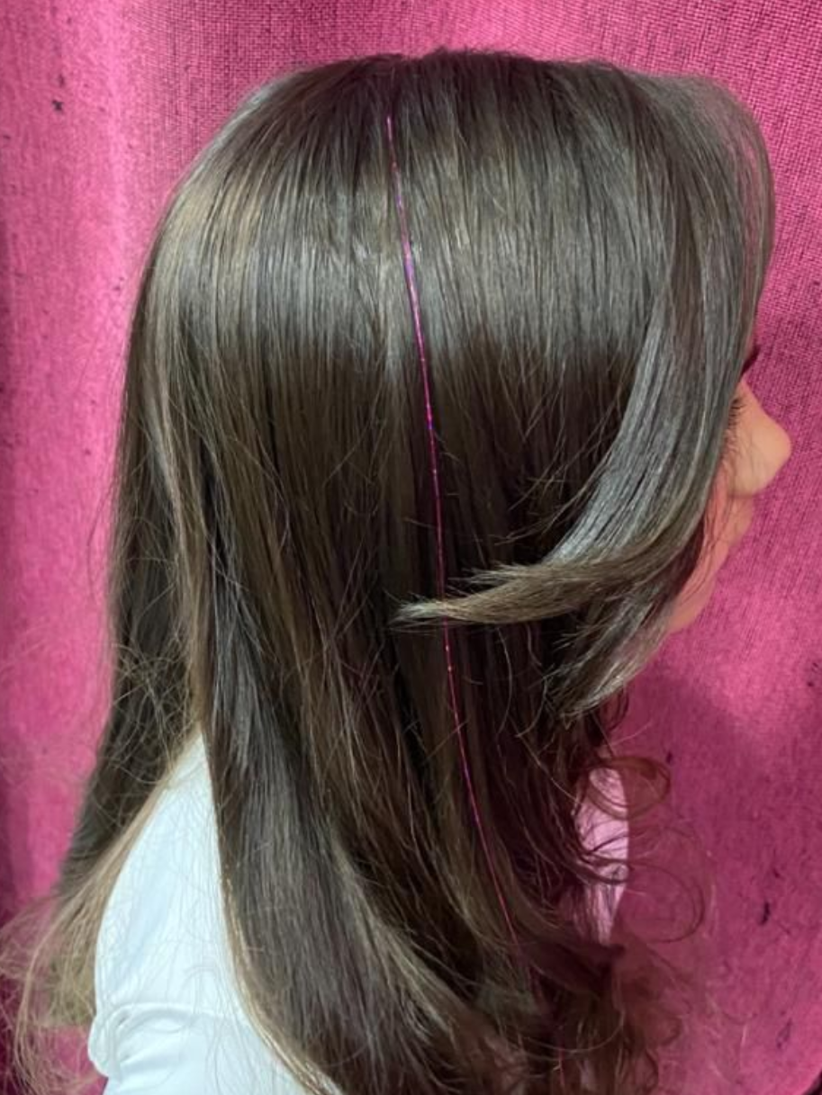 Chica con pelo castaño largo en capas, vestida con una camisa blanca, con mechones de oropel rosados, sobre un fondo rosa.