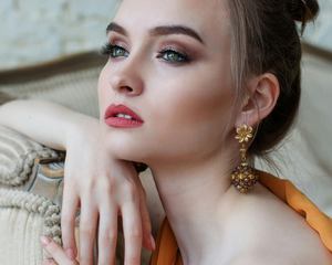 Mujer con maquillaje, con pendientes, apoyada en un sofá, mirando hacia arriba con una leve sonrisa.
