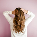 Mujer con cabello castaño largo y ondulado, con las manos en el cabello, sobre un fondo rosa y vestida con una sudadera blanca.