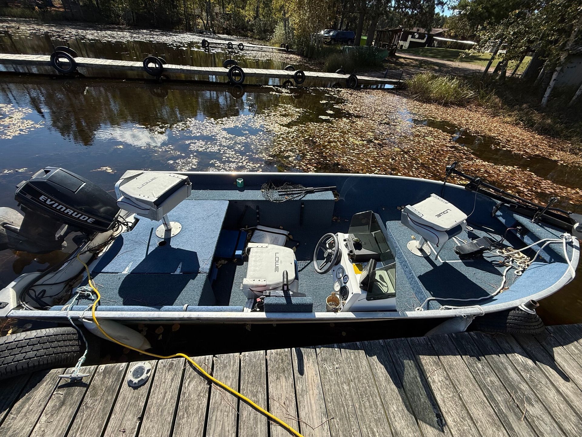 Boat docked at a wooden pier. Blue interior, black motor, surrounded by water and fall foliage.