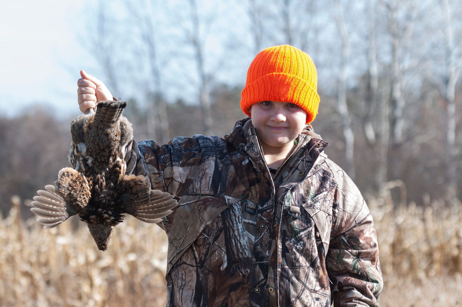 Person in camouflage holds up a bird they have hunted, orange hat, outdoors.