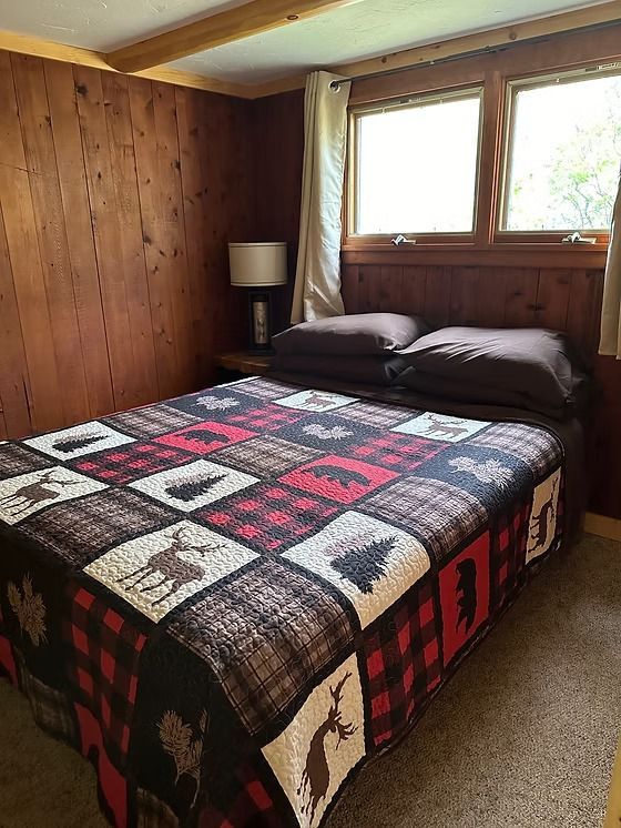 Bedroom with a rustic quilt, wooden paneling, and two windows.