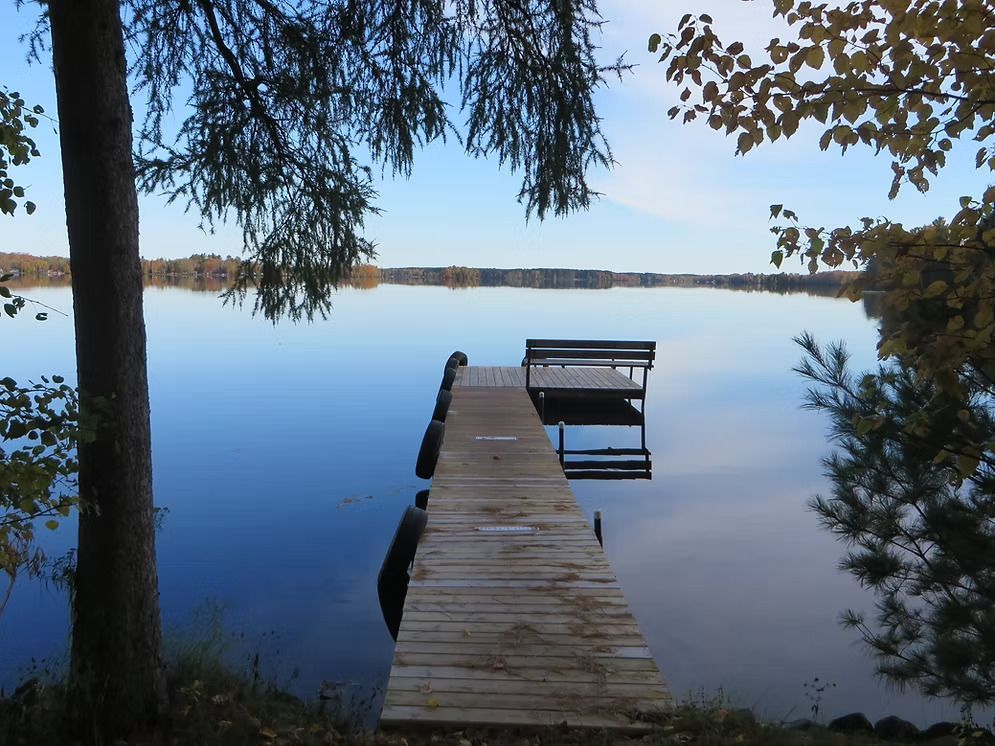 Wooden dock with bench extending into a calm lake under a clear sky. Autumn trees frame the view.