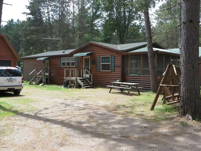 Log cabin with screened porch, picnic table, and swing set in a wooded area. A white vehicle is parked.