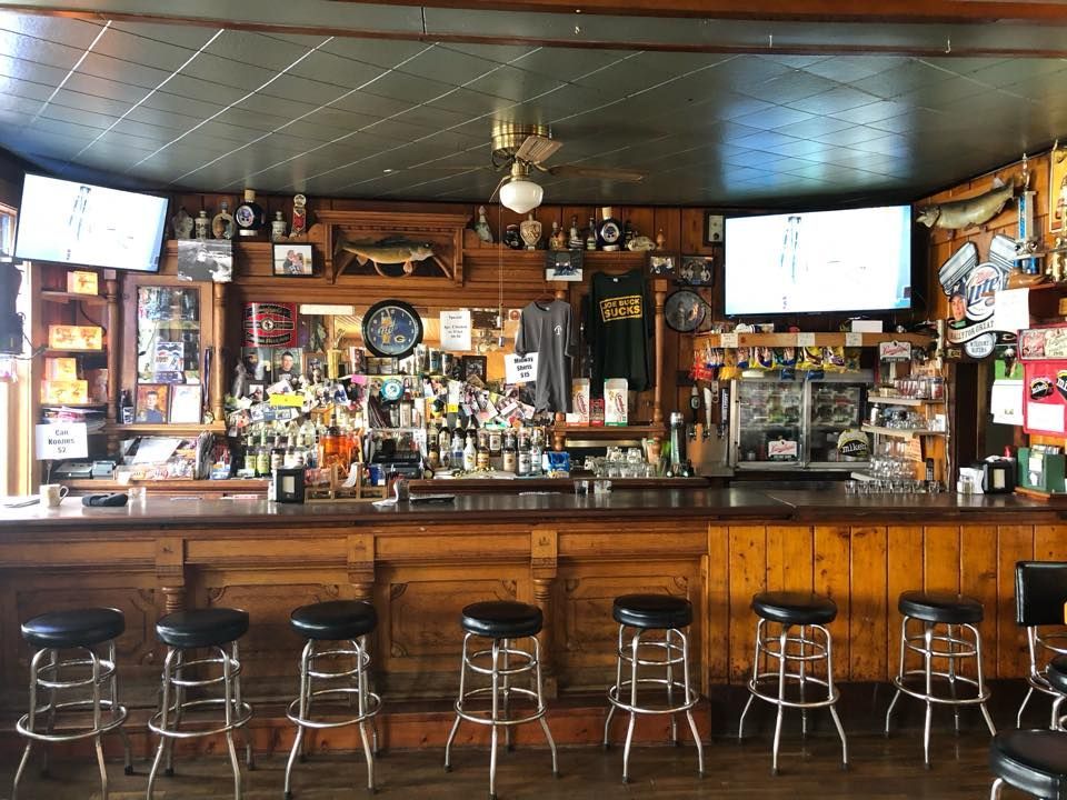 A wooden bar with stools, drinks, bottles, and TVs. The bar has various decorations on a wood-paneled wall.