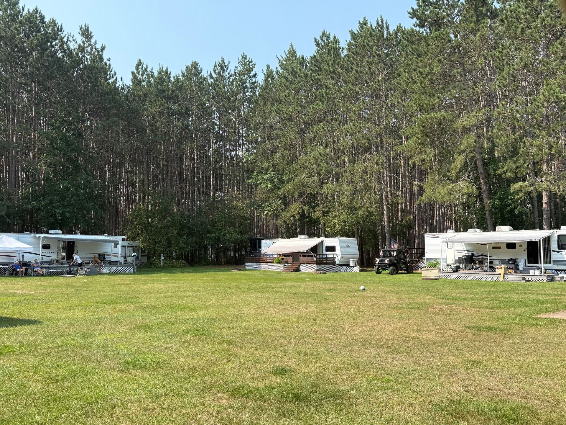 Campground with three RVs on grassy area, tall trees in the background. Sunny, daytime.