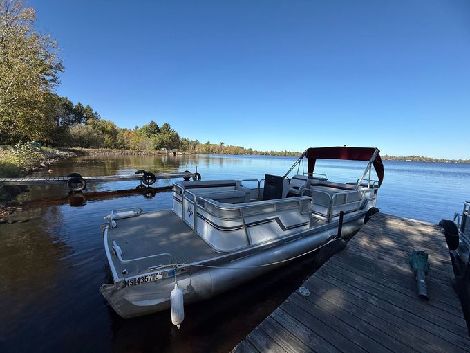 Pontoon boat docked on a wooden pier, calm lake, clear blue sky, trees in the background.