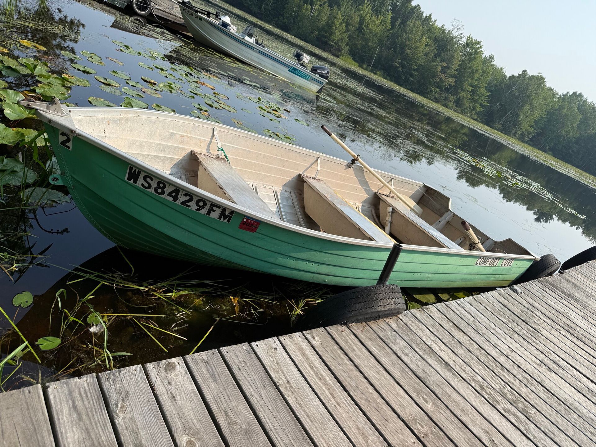 Green rowboat at a wooden dock, with another boat in the background on a calm lake.