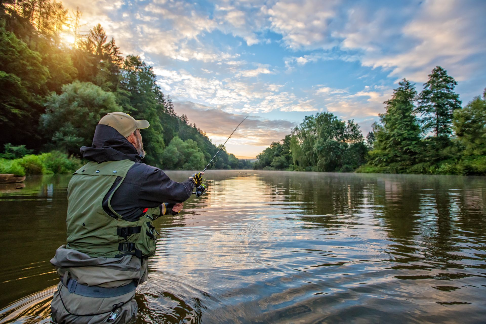 A person wearing a fishing vest and cap casts a line into a calm, sunlit river bordered by trees at dawn.