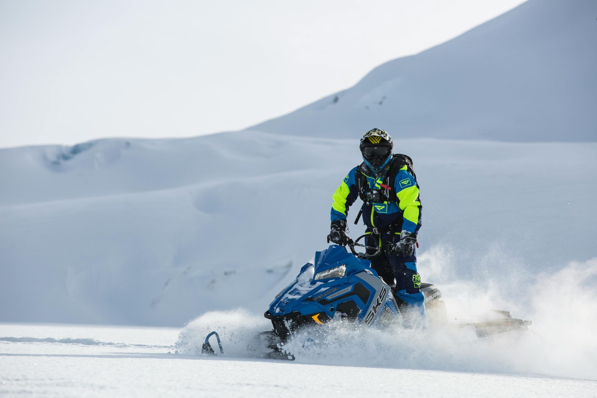 Snowmobiler riding across a snowy landscape, kicking up snow.