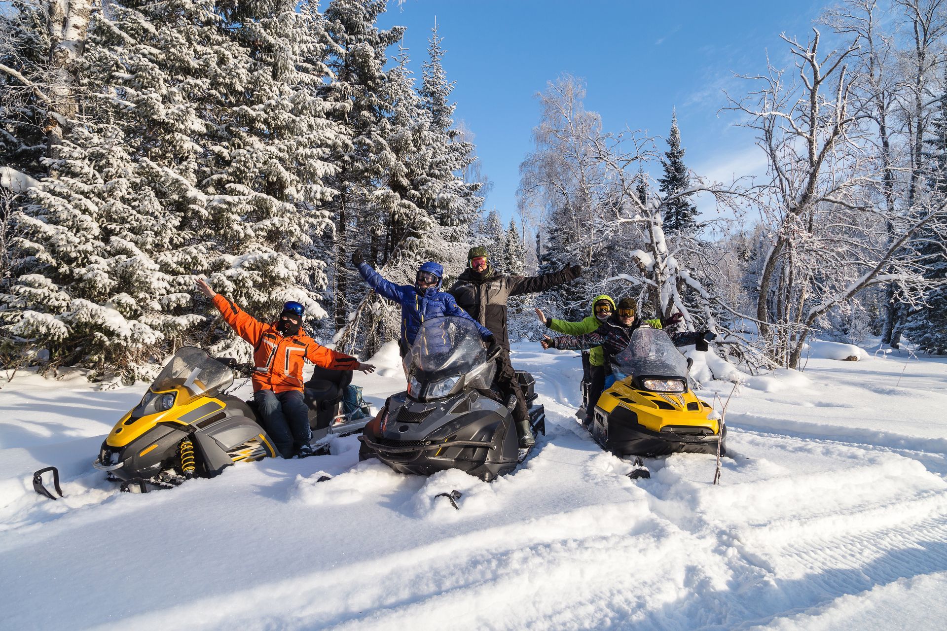 Four snowmobilers in bright gear posing in a snowy forest under a blue sky