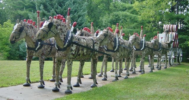 Sculpture of a carriage pulled by ten decorated horses on a path, in a grassy area with trees.