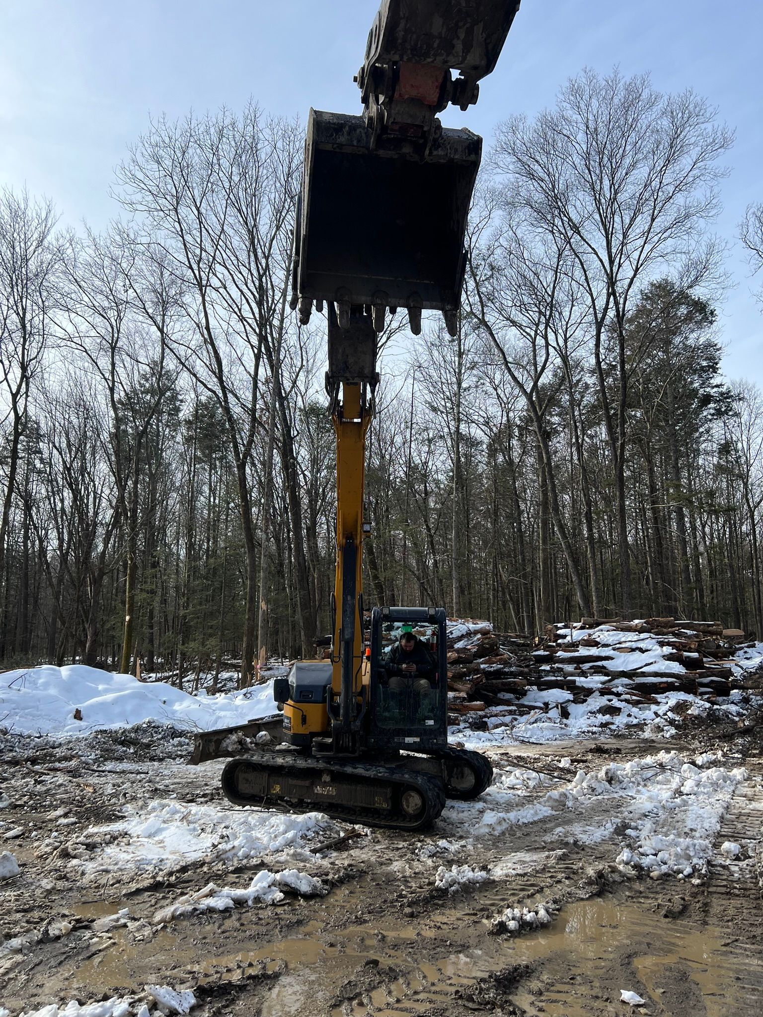 Yellow excavator with raised bucket in a snowy forest clearing.
