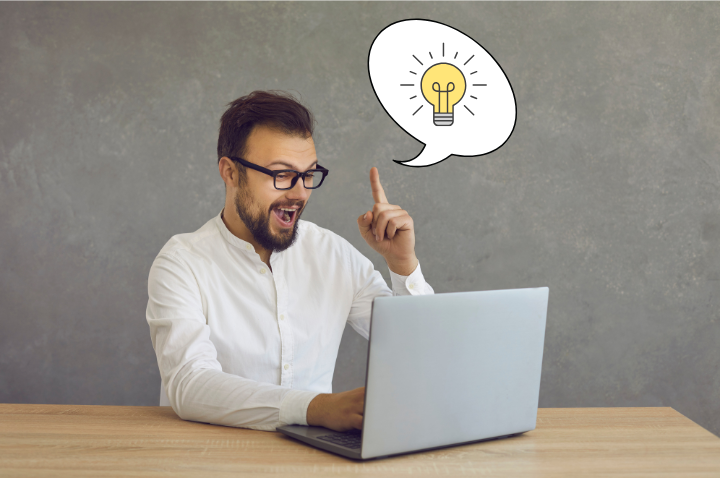 Man with glasses excitedly points upward, a lightbulb icon in a speech bubble above a laptop on a wooden desk.