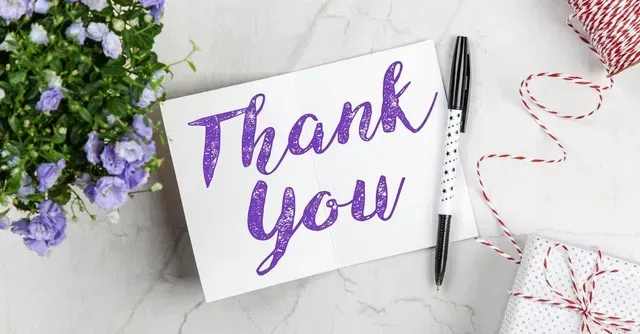 Thank you card with purple script, next to a pen, a small gift box, a ball of twine, and purple flowers.