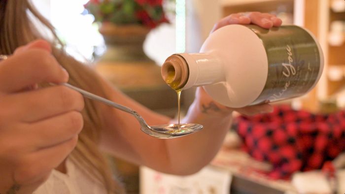 A woman is pouring olive oil into a spoon from a bottle.