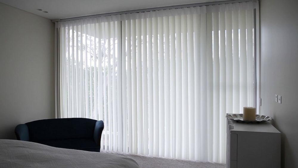 A Bedroom With a Bed, Chair and Sliding Glass Doors With Vertical Blinds — Whitsunday Blinds & Security in Airlie Beach, QLD