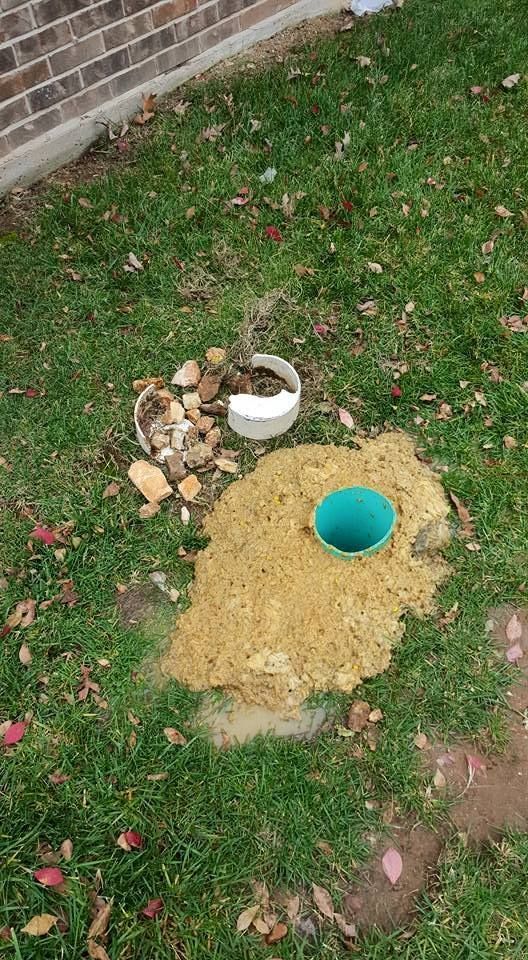Green pipe in tan dirt on grass, next to a white pipe with rocks. Brick wall in the background.