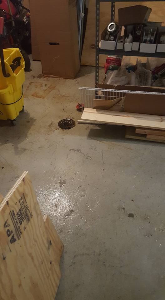 Concrete floor in a cluttered garage, with a drain, wood, and a yellow container visible.