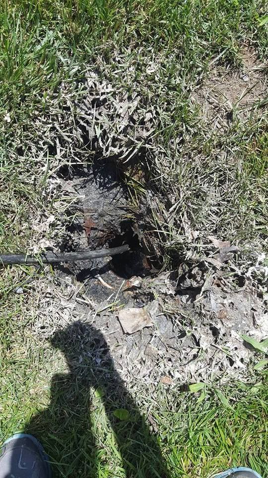 Charred debris and wire in a patch of dead grass, viewed from above. Shadow of person's legs.
