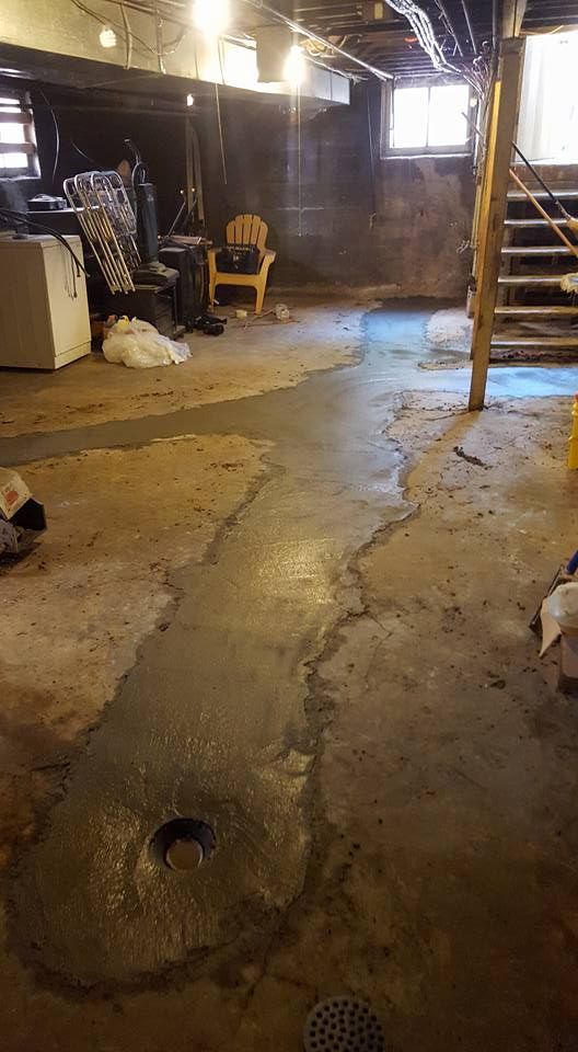 Basement floor with a drain and water. Concrete is cracked and shows water damage. Staircase and furniture visible.