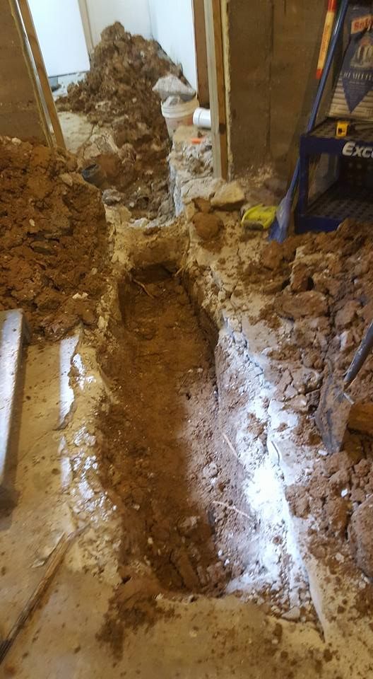 Trench dug through a concrete floor, surrounded by dirt and debris, near a doorway.
