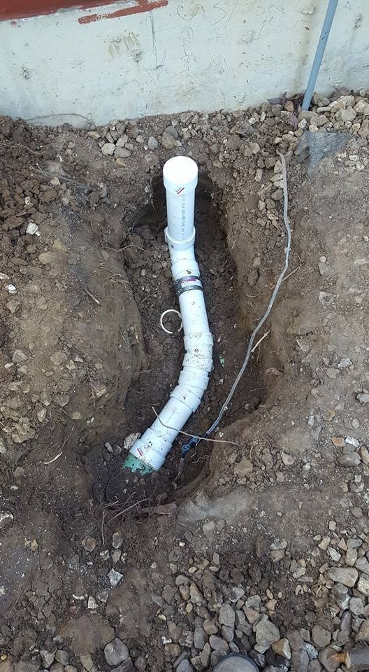 A partly buried white PVC pipe extends from a foundation wall, with a flexible section and a small wire visible.