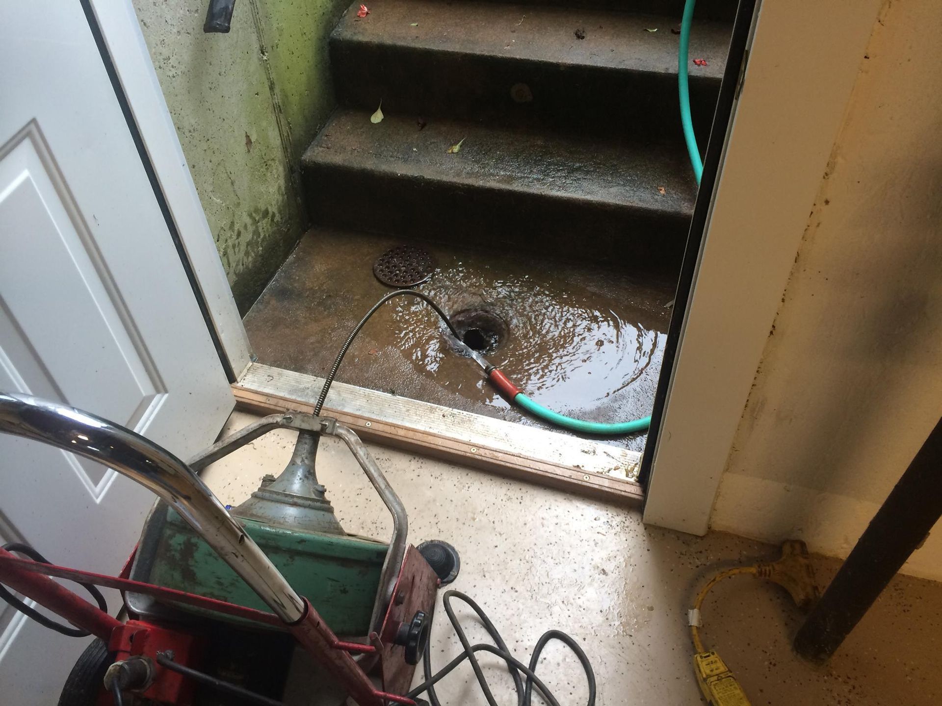Sewer snake clears a drain in a basement stairwell filled with water; a door stands open nearby.
