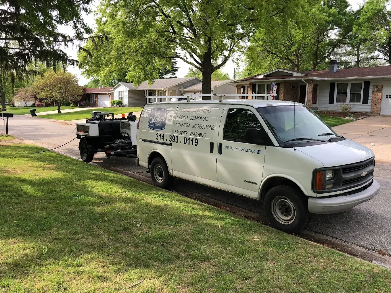 White van with trailer parked on a residential street. The van has business information on the side.