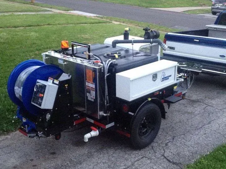 Road maintenance trailer with a blue hose reel, connected to a truck parked on a cracked driveway.