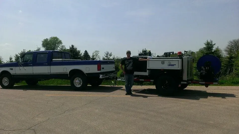 Blue and white pickup truck towing a trailer with a man standing beside it on a sunny day.