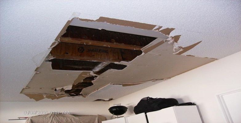 Damaged ceiling with a large hole revealing wooden beams. White walls surround the destruction.