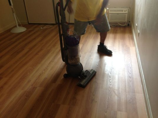 Person vacuuming a room with wood-look flooring; the person wears shorts and a yellow shirt.