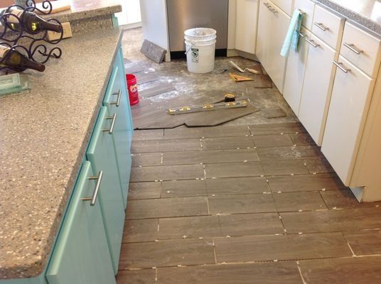 Kitchen undergoing tile flooring installation: gray wood-look tiles, white cabinets, turquoise cabinets, bucket, tools.