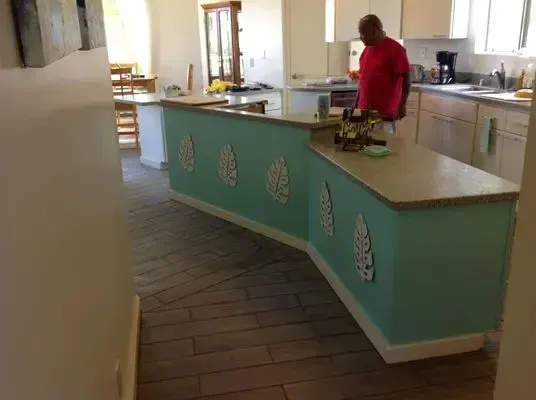 Kitchen with turquoise island, decorative leaves, wooden floor, and a person in a red shirt.