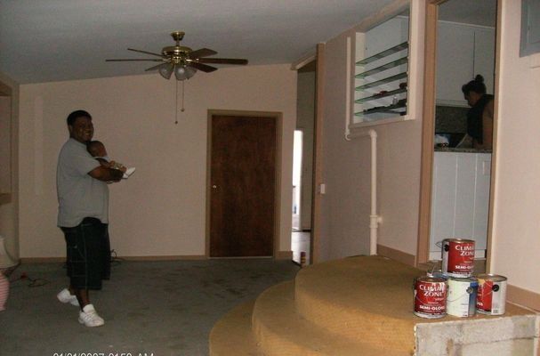 Man holding a baby in a living room; brown door, pink walls, and a woman in the doorway.