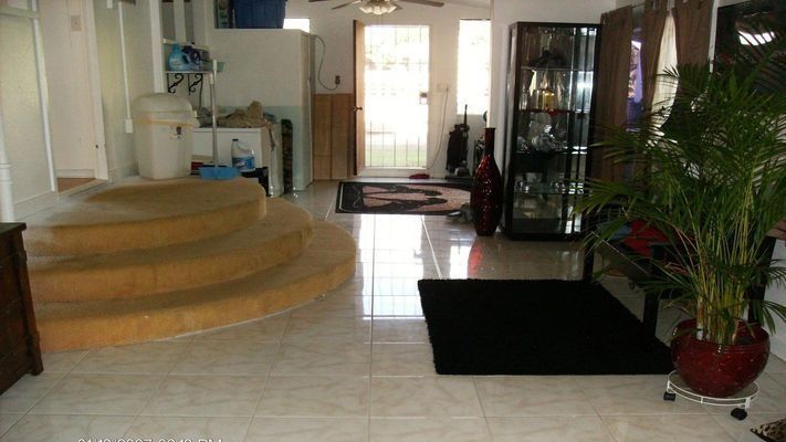A living room with tile floor, sunken seating area, and potted plant. Bright sunlight shines through the front door.